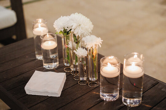 Lit candles in transparent glass jars on a wooden table A glass vases of water with burning candles inside on the table Wedding table set decor Vases with white flowers Cropped photo Close up	