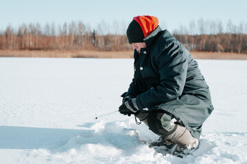 Fototapeta na wymiar Winter fishing, side view of caucasian male fisherman sitting with rod on frozen water near drilled hole in ice on sunny day, outdoors. Hobby and sports, active leisure and recreation concept