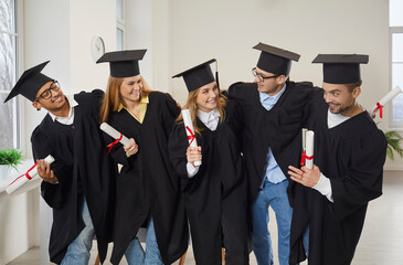 Diverse group of happy university students celebrating graduation. Cheerful, smiling, optimistic young classmates and friends in graduate academic hats and gowns hugging and having fun in classroom