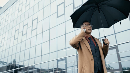 Mature businessman holding umbrella and talking on mobile phone on urban street.