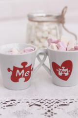 Two valentine's coffee mugs with red hearts and words you and me, Marshmallows in a glass on white background close up, side view