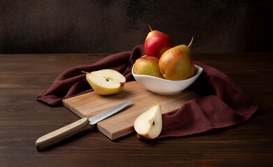 Still life with Trout pears. Several fruits lie in a white bowl standing on a wooden chopping board. Next to the bowl is a sliced pear and a knife. Low key, brown background, horizontal orientation.
