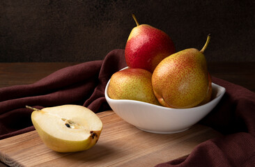 Trout pears in a white porcelain bowl standing on a wooden board. Next to the bowl is half a sliced pear. Brown background, close-up, horizontal orientation.