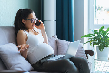 Pregnant woman with glasses sit on sofa in living room and talk lively on mobile phone putting...