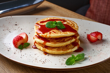 Stack of pancakes with of strawberry slices and caramel syrup on a wooden table.