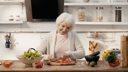happy senior woman cutting bell pepper near fresh cherry tomatoes, lettuce and carrots.