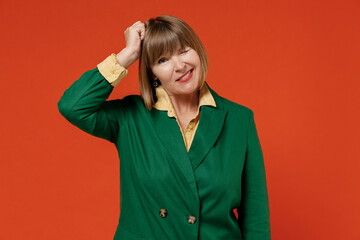 Elderly mistaken puzzled sad confused caucasian woman 50s wearing green classic suit scratch holding head isolated on plain orange color background studio portrait. People business lifestyle concept.