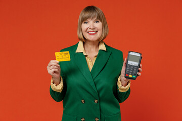 Elderly smiling fun woman 50s wearing green classic suit hold wireless modern bank payment terminal to process acquire credit card payments isolated on plain orange color background studio portrait