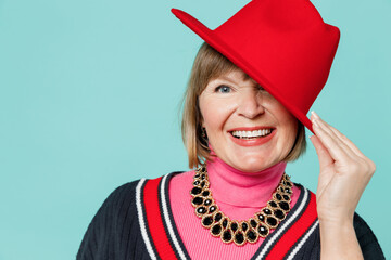 Close up elderly smiling happy cheerful beautiful stylish cool fashionable woman 50s in necklace shirt cover eye with red hat isolated on plain pastel light blue background. People lifestyle concept.