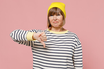 Elderly disappointed displeased sad woman 50s wear striped shirt yellow hat showing thumb down dislike gesture isolated on plain pastel light pink background studio portrait. People lifestyle concept.