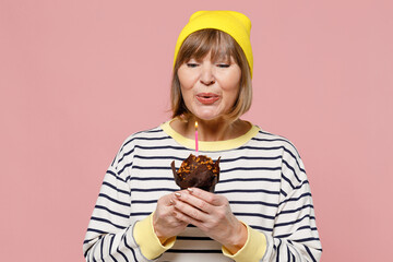 Elderly caucasian stylish woman 50s wearing striped shirt yellow hat hold sweet birthday cake blow...