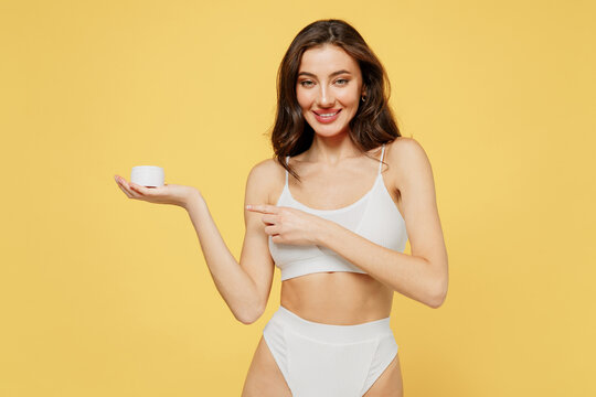Smiling lovely attractive young brunette woman 20s in white underwear with perfect fit body point finger on container of moisturizer cream look camera isolated on plain yellow color background studio.