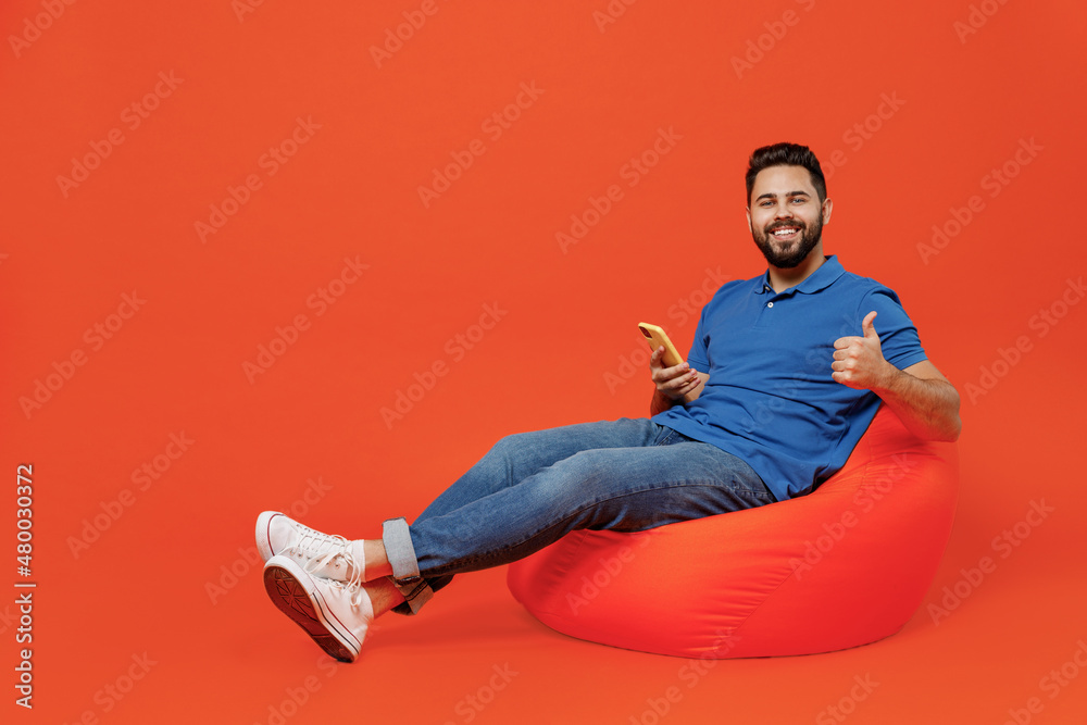 Poster full body young smiling happy man wear blue t-shirt sit in bag chair hold in hand using mobile cell 