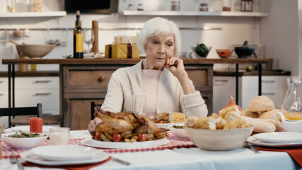 upset senior woman sitting alone near thanksgiving dinner on table in kitchen.