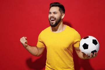 Fototapeta Jubilant fun young bearded man football fan in yellow t-shirt cheer up support favorite team hold soccer ball look aside clenching fists say yes isolated on plain dark red background studio portrait. obraz