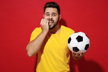 Worried vivid young bearded man football fan in yellow t-shirt cheer up support favorite team hold in hand soccer ball look camera biting nails isolated on plain dark red background studio portrait.