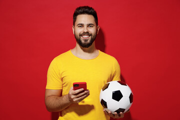 Smiling laughing beautiful young bearded man football fan in yellow t-shirt cheer up support favorite team hold soccer ball use mobile cell phone isolated on plain dark red background studio portrait.