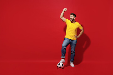 Fototapeta na wymiar Full size body length young bearded man football fan in yellow t-shirt cheer up support favorite team hold foot on soccer ball do winner gesture isolated on plain dark red background studio portrait.