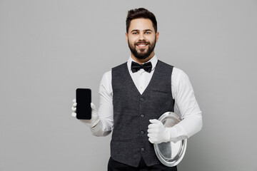 Smiling young barista male waiter butler man 20s wear white shirt vest elegant uniform work at cafe hold mobile phone with blank screen isolated on plain grey background. Restaurant employee concept.