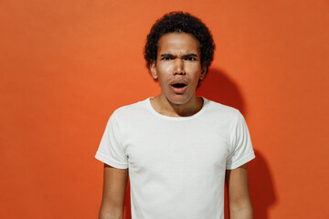 Shocked surprised impressed frightened upset young black curly man 20s years old wears white t-shirt looking camera keeping mouth wide open isolated on plain pastel orange background studio portrait.