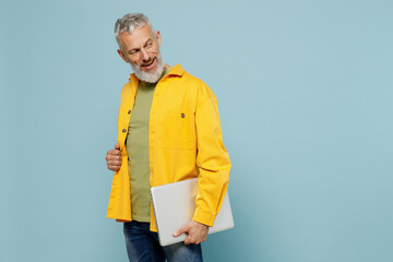 Elderly side view happy gray-haired mustache bearded man 50s in yellow shirt hold closed laptop pc computer look aside on workspace copy space isolated on plain pastel light blue background studio