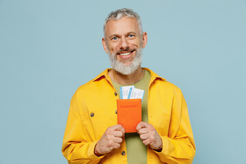 Traveler tourist elderly gray-haired bearded man in yellow shirt hold passport ticket isolated on plain blue color background. Passenger travel abroad on weekends getaway. Air flight journey concept.