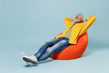 Full body sleeping elderly gray-haired mustache bearded man 50s in yellow shirt sit in bag chair hold hands behind neck isolated on plain pastel light blue background studio. People lifestyle concept.