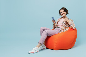 Full body young smiling cheerful fun happy woman 20s in brown shirt sit in bag chair hold use mobile cell phone show thumb up gesture isolated on pastel plain light blue background studio portrait