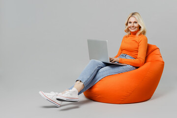Full body elderly smiling happy blonde woman 50s in orange turtleneck sit in bag chair hold use...