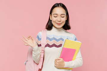 Teen student girl of Asian ethnicity in sweater backpack hold books hold hands in yoga om aum gesture meditate isolated on pastel plain light pink background Education in university college concept