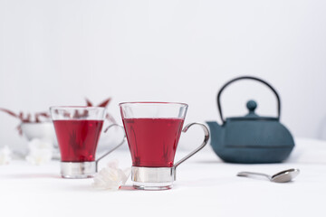 
Dos tazas transparentes de té de bayas con tetera de hierro azul sobre una mesa blanca y un fondo gris claro