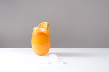 Jugo de naranja exprimido en un vaso de cristal con rodaja de naranja y hielo sobre una mesa blanca con un fondo gris	