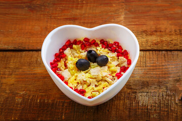 Puff salad with chicken breast and pineapple, garnished with pomegranate in a heart salad bowl on a wooden table.