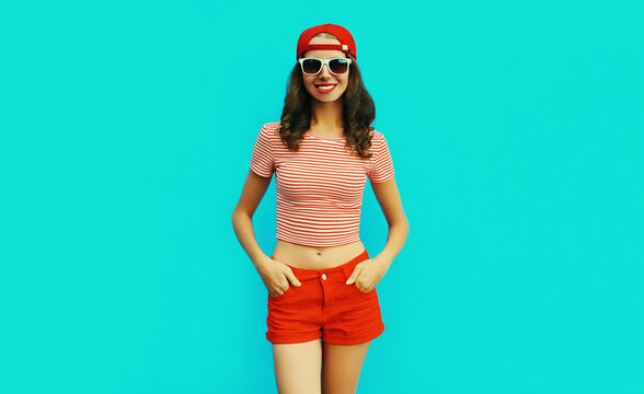 Summer portrait of happy smiling young woman wearing baseball cap, red shorts on blue background