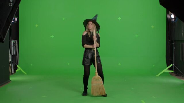 Young caucasian woman wearing witch hat for halloween parties over green screen background. Girl having fun with a broom making like playing the guitar . Halloween concepte. Chroma key. 4k video