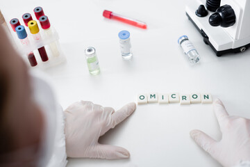 partial view of blurred scientist near cubes with omicron lettering, vaccine vials and test tubes in lab.