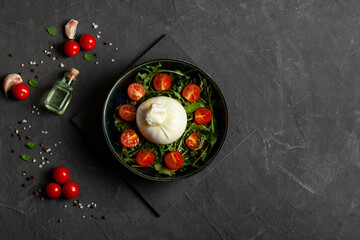 Salad with traditional italian burrata cheese with arugula and tomatoes om dark concrete table with cooking ingredients cherry tomatoes, basil, garlic and olive oil. Top view. Copy space.