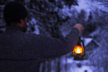 A man with a kerosene lamp in his hand walks through the snowy forest at night to his house in the village