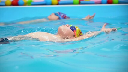 Two young Caucasian boys with goggles exercising freestyle swimming on back and breathing in pool with blue water. Indoor view. Slow motion. Activity sport.