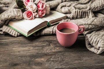 Morning coffee. A cup of coffee on a wooden table, an open book and a warm sweater against the background of a bouquet of flowers. Cozy morning.