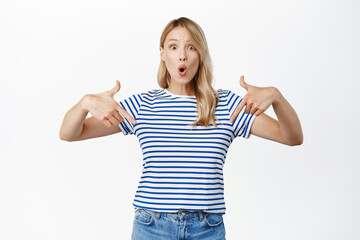 Fototapeta na wymiar Impressed blond girl shows advertisement, points fingers down and looks amazed, demonstrates awesome sale in store, click link, pointing at banner, white background
