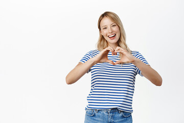 Happy beautiful girl laughing and smiling, showing heart gesture, i love like, standing in t-shirt...