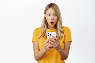 Girl looks surprised and shocked at mobile phone, woman reading message with amazed face expression, standing in yellow t-shirt over white background