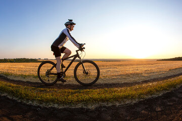 The cyclist rides a bike on the road near the field against the backdrop of the setting sun. Outdoor sports. Healthy lifestyle.