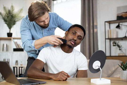 Portrait of stylish African man looking into the camera while his Caucasian friend shaving his neck with an electric razor to create a stylish hairstyle. Multiracial gay couple