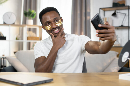 Close-up of a young positive African guy in a white t-shirt uses patches under his eyes while making selfie on smartphone. The concept of healthy skin, hygiene and health