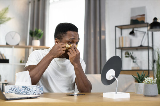 The concept of modern life, hygiene and beauty. Sporty happy African guy applying golden patches under his eyes looking in the mirror sitting at home in a modern room