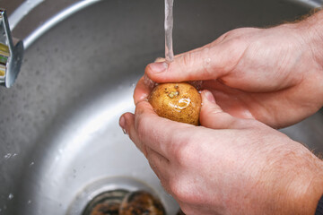 A man washes raw potatoes in a sink under water to bake them in foil in the oven.