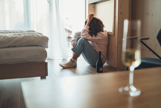 Sad woman sitting on the floor in the bedroom with a bottle of alcohol. Unfocused wine glass on the foreground table. Mental health and alcoholism problems concept image..