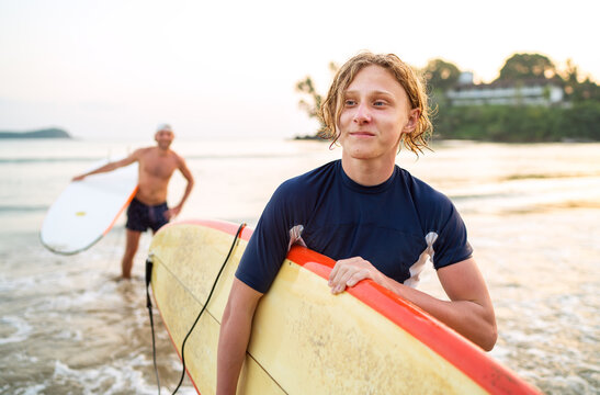 Portrait of a teen boy with a surfboard after surfing with his father. They are smiling and walking out of the water. Family active vacation concept.